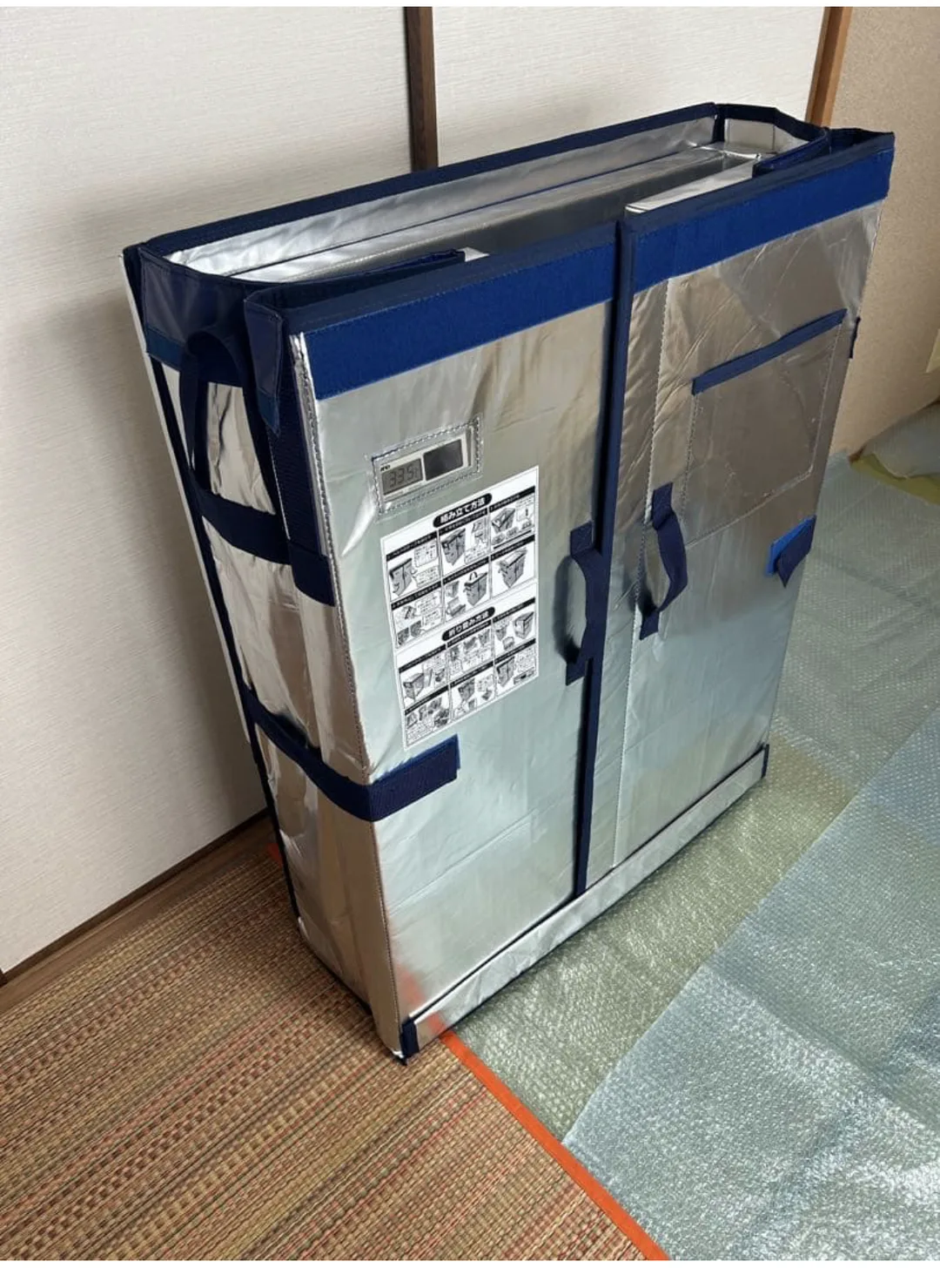 同じ保冷ボックスを折り畳んだ状態。厚さが10分の1ほどの板状になり、紺色のベルトで留められて壁際に立てかけられている。前面のデジタル温度計は33.5℃を表示している。