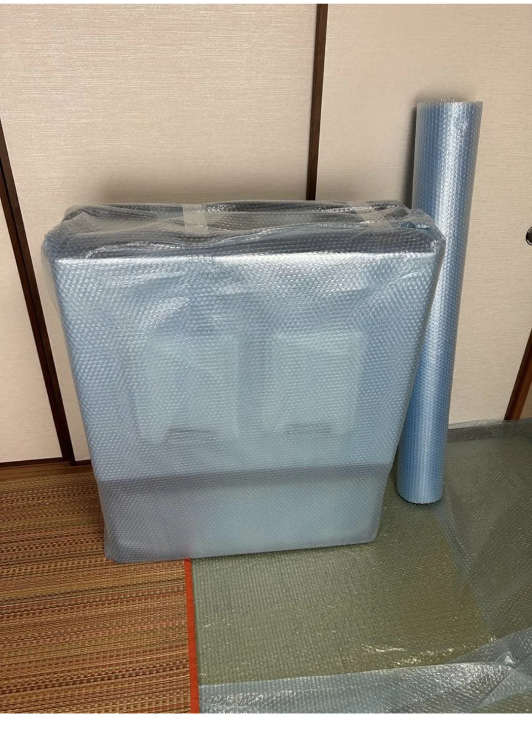 プチプチで全体を包み終えた折りたたみ状態の保冷ボックスが壁際に立てられ、その右隣にプチプチのロールが立てて置かれている。
