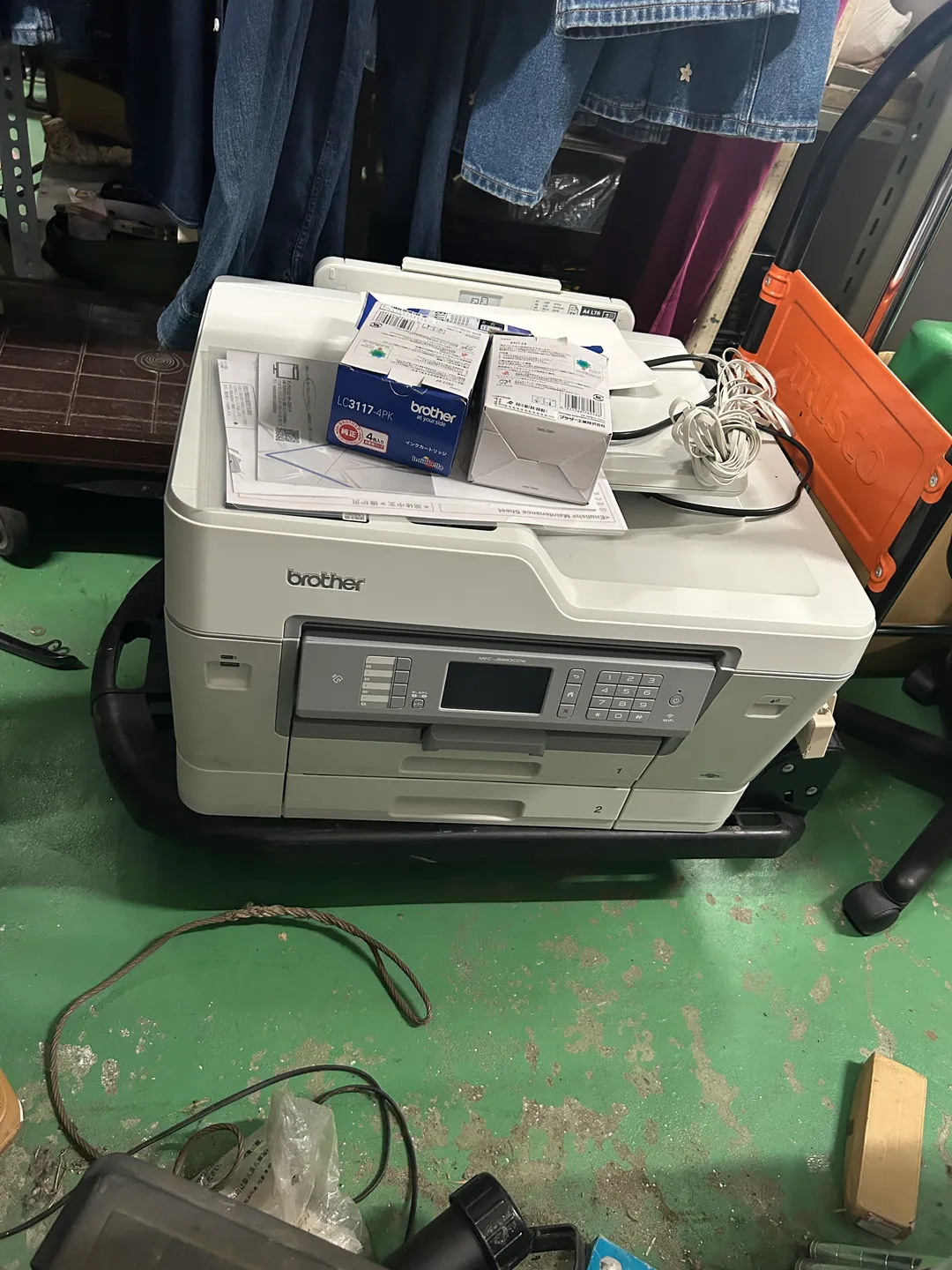 倉庫の作業台に置かれたブラザーのA3インクジェット複合機 MFC-J6983CDW。天板の上に「LC3117-4PK インクカートリッジ 4個入」の箱と、ブラザー純正の白い部品箱、青いパッケージ、取扱説明書やメンテナンスシート等の書類一式、電源コードがまとめて載っている。