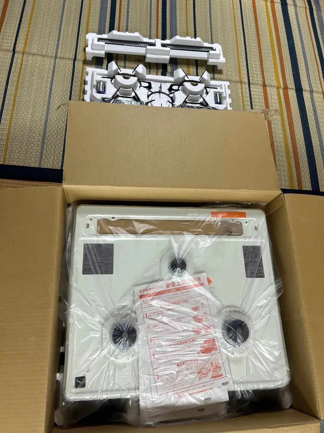 段ボール箱から取り出した本体。白いホーロー天板がビニールに包まれ、3つのバーナー穴が見えている。天板の上に「安全上のご注意」と書かれた赤い注意書きの紙が置かれ、奥の緩衝材には五徳とグリル部品が並んでいる。