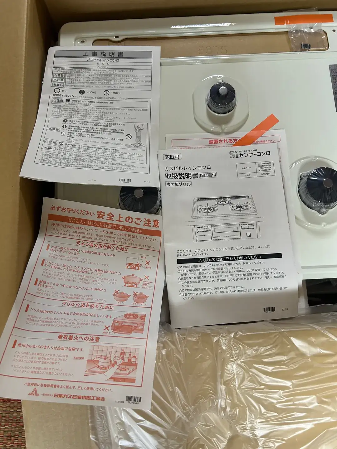 天板の上に広げた3種類の書類。左上に「工事説明書 ガスビルトインコンロ」、左下に「必ずお守りください 安全上のご注意」（日本ガス石油機器工業会）、右に「家庭用 ガスビルトインコンロ 取扱説明書 保証書付 片面焼グリル」の冊子が置かれている。奥にはオレンジ色の帯が付いた「設置される方へ」の紙も見えている。