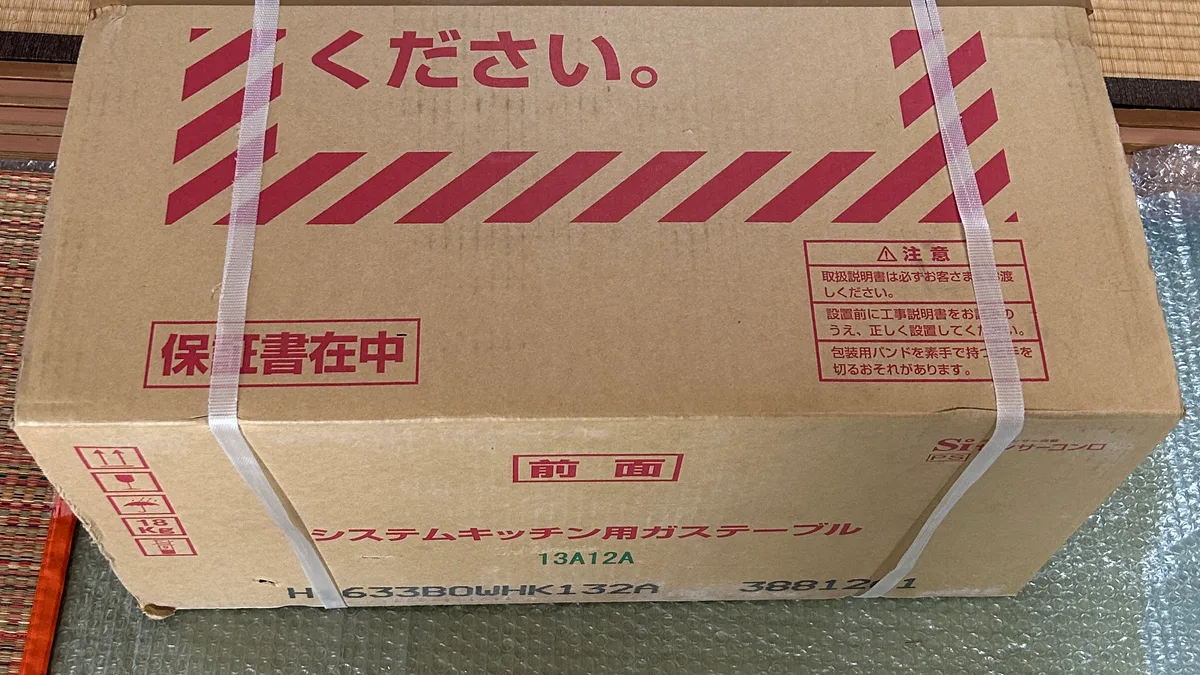 段ボール箱の側面。「保証書在中」の赤枠、「前面」の表示、「システムキッチン用ガステーブル」「13A12A」「H1633B0WHK132A」の印刷が並び、右下に「Si センサーコンロ」のロゴ。左端に重量18kgを示すマークが見える。梱包バンドが2本かかっている。