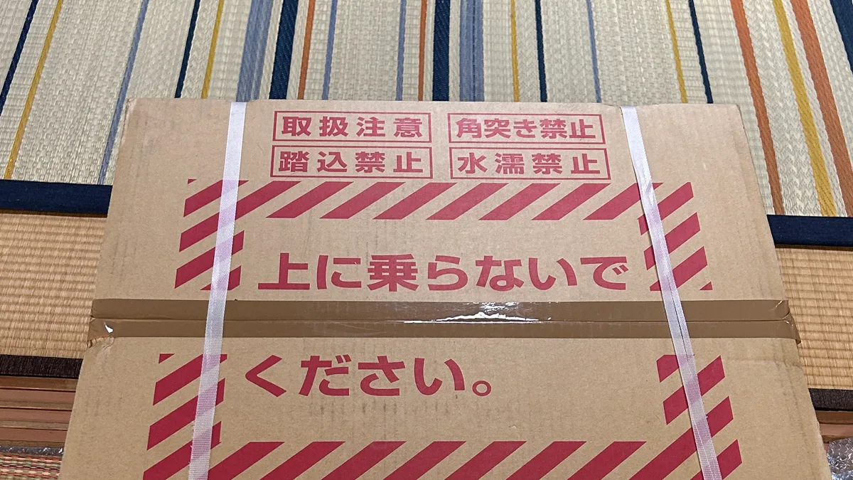 段ボール箱の上面。「取扱注意」「角突き禁止」「踏込禁止」「水濡禁止」の赤い枠と、斜線に囲まれた「上に乗らないでください。」の大きな印刷。梱包バンドが2本かかっている。