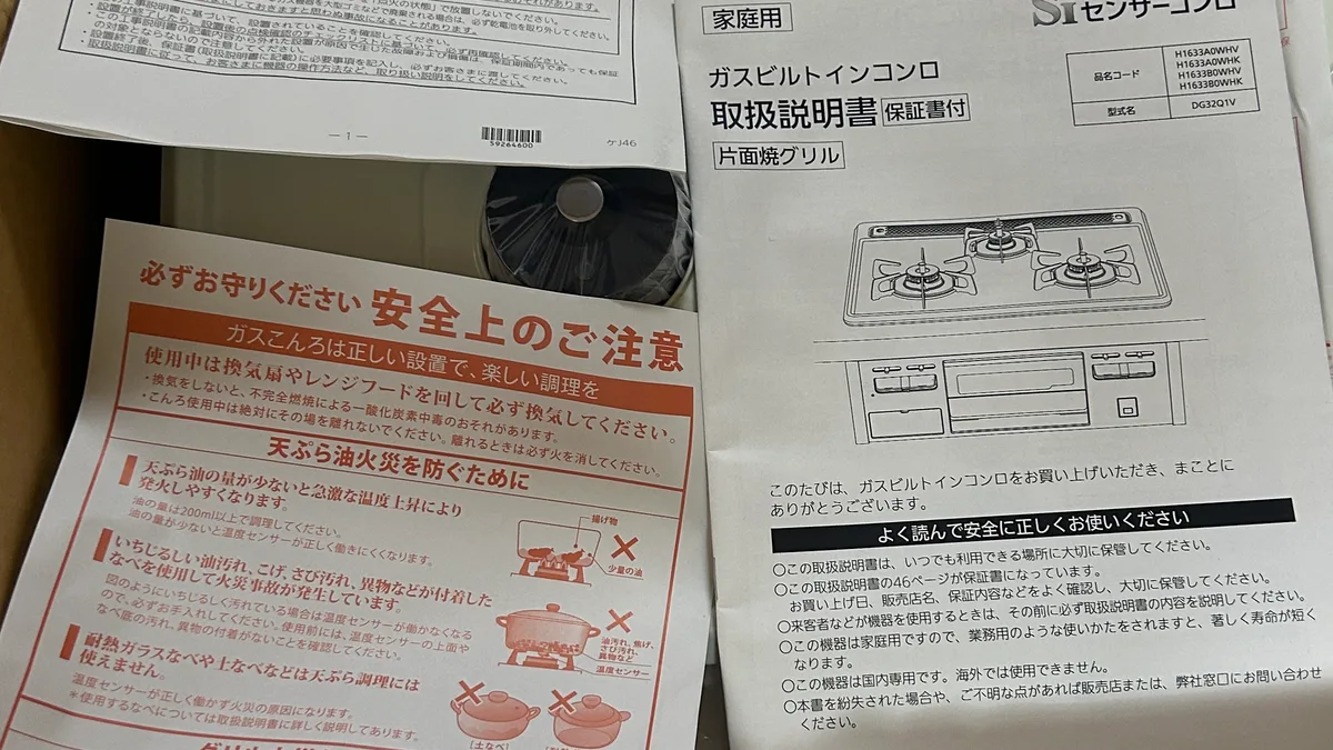 天板の上に広げた書類。左上に「工事説明書 ガスビルトインコンロ」、左下に「必ずお守りください 安全上のご注意」（ガスこんろは正しい設置で、楽しい調理を／天ぷら油火災を防ぐために）、右に「家庭用 ガスビルトインコンロ 取扱説明書 保証書付 片面焼グリル」の冊子。取扱説明書の表紙には品名コードと型式名の表が印刷されている。