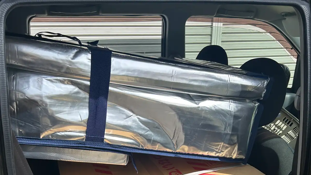 車の後部座席に積み込まれた銀色の保冷ボックスと、その下に敷かれた大型の段ボール箱。車のドアが開いた状態で、屋外の駐車場から車内を撮影している。