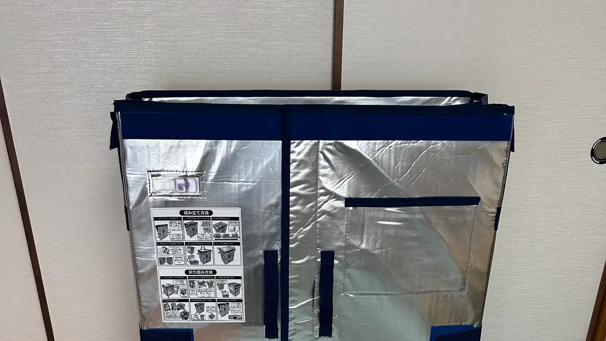 ふすまの前に置かれた、銀色の断熱シート製で紺色のベルトが付いた折りたたみ式の大型保冷ボックス。前面に組み立て方法と折り畳み方法のイラスト入り説明シールが貼られている。