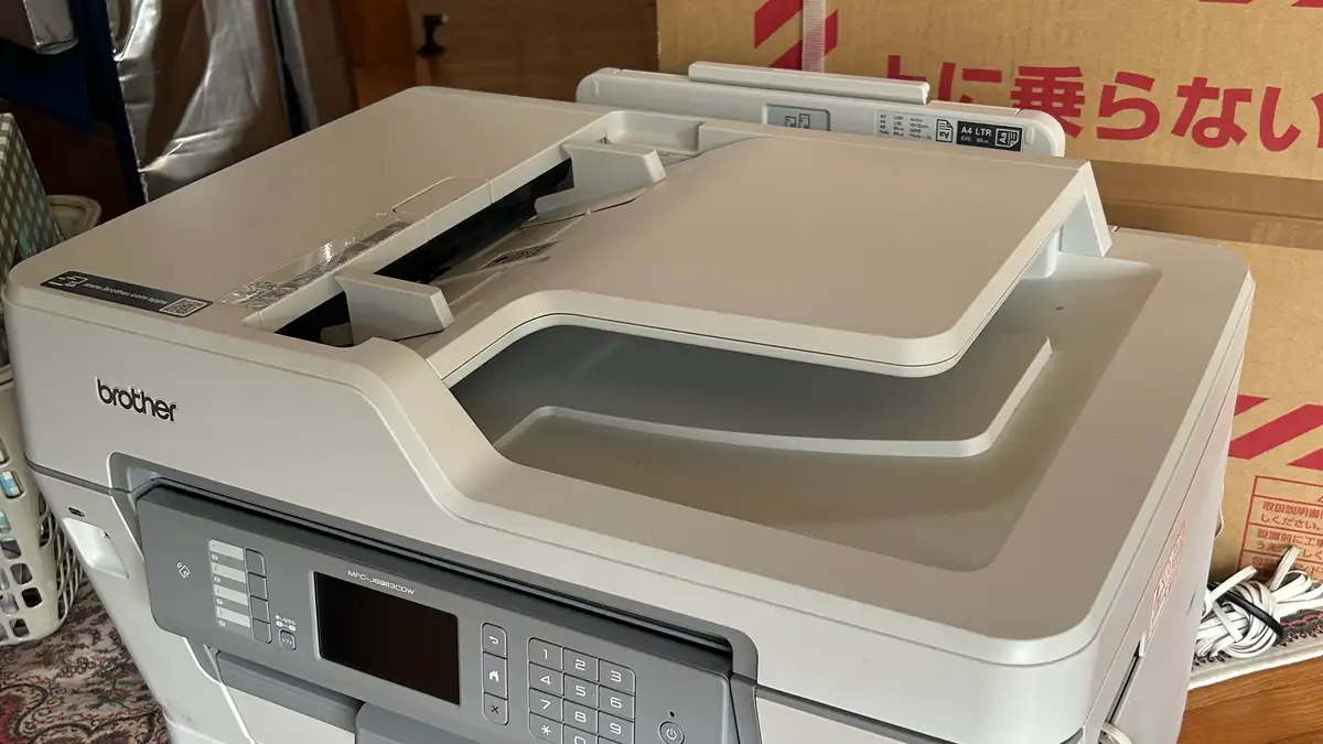 ブラザー工業のA3インクジェット複合機 MFC-J6983CDW を斜め上から撮った写真。白い本体の操作パネルに型番が印字され、上部の原稿カバーとADFトレイが開いた状態。奥には赤い印刷の入った大型段ボール箱が置かれている。